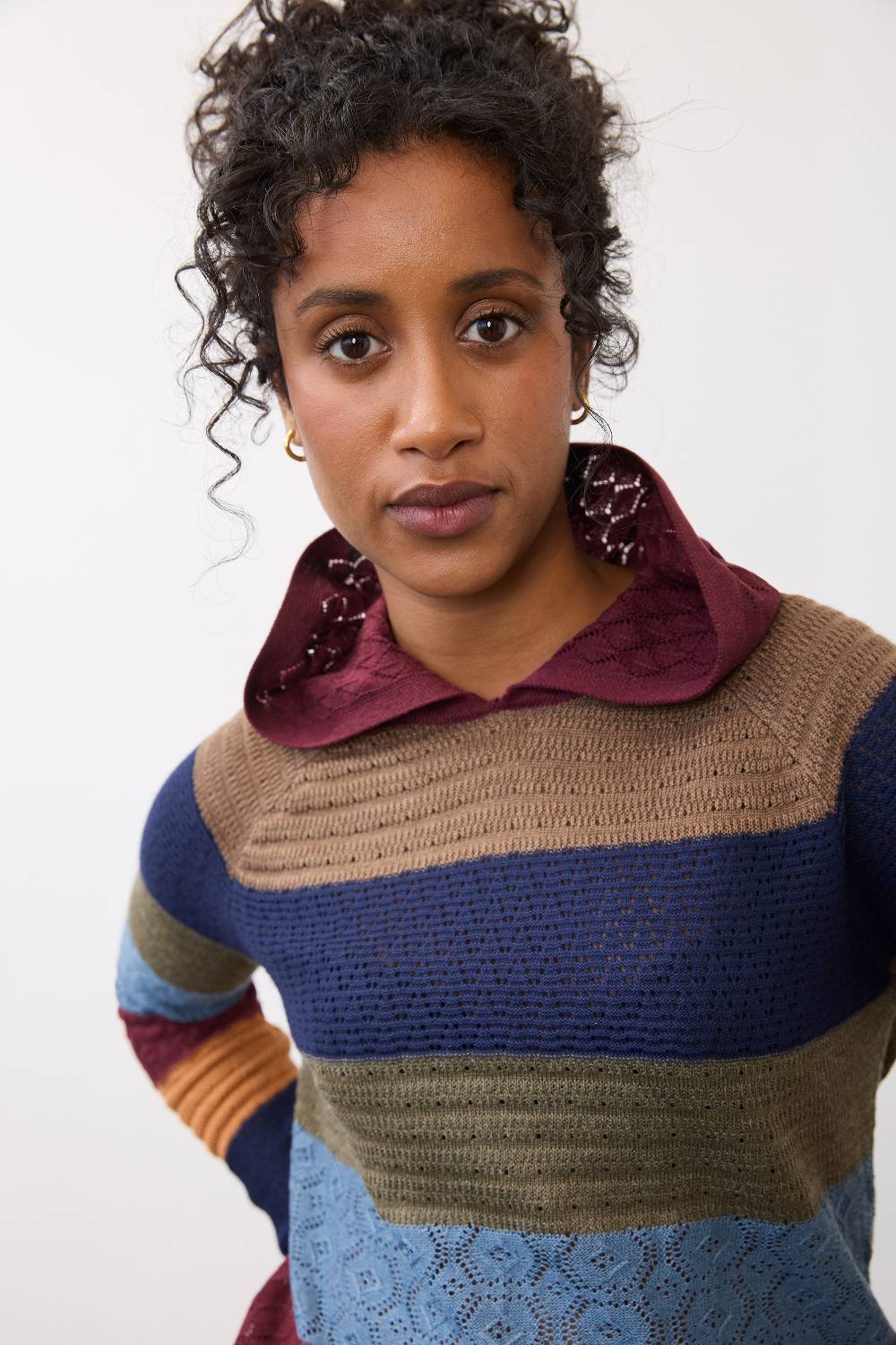 Woman wearing a multi coloured striped knit hoodie against a light backdrop.