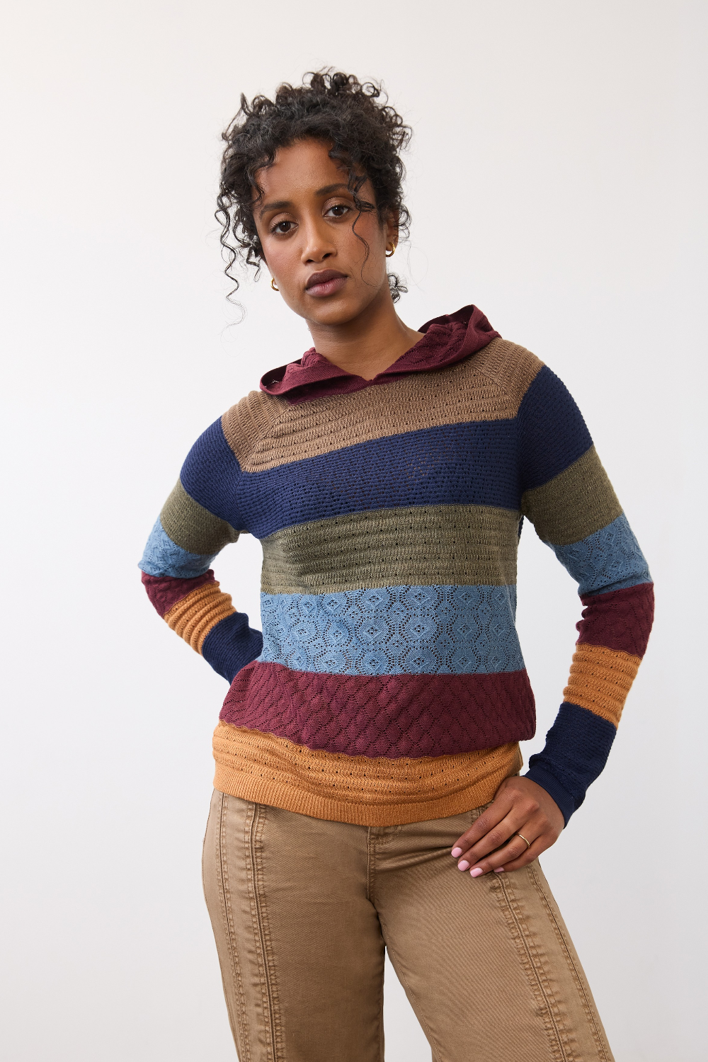 Woman wearing a multi coloured striped knit hoodie with tan pants against a light backdrop.