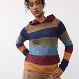 Woman wearing a multi coloured striped knit hoodie with tan pants against a light backdrop.