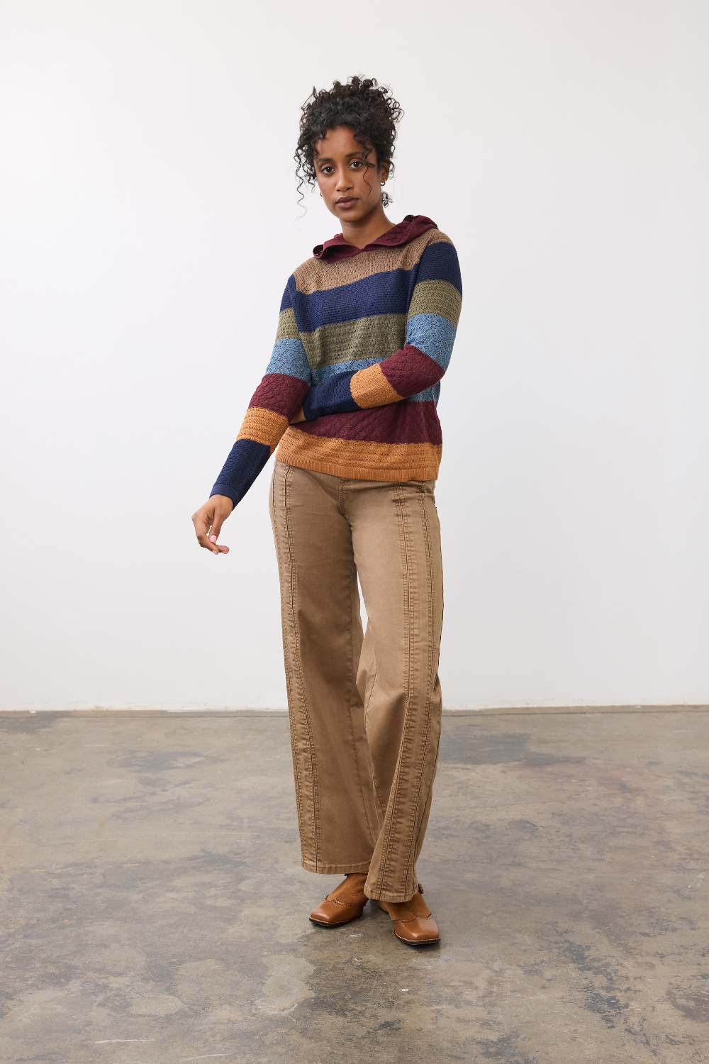 Woman wearing a multi coloured striped knit hoodie with tan pants and light brown shoes against a light backdrop.