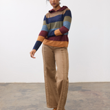 Woman wearing a multi coloured striped knit hoodie with tan pants and light brown shoes against a light backdrop.