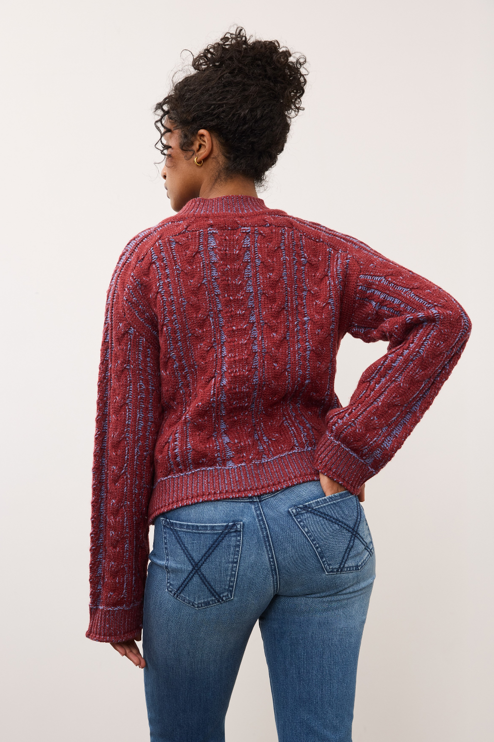 Woman, shown from the back, wearing a red and blue mix cable knit crew neck sweater with blue jeans against a light studio backdrop.