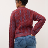 Woman, shown from the back, wearing a red and blue mix cable knit crew neck sweater with blue jeans against a light studio backdrop.