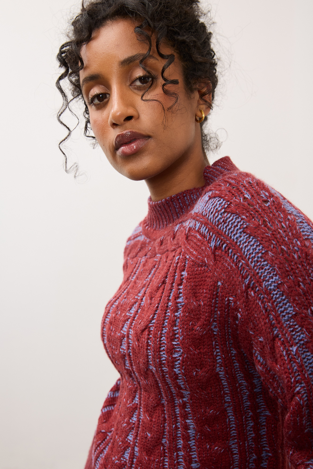 Close up of a woman wearing a red and blue mix cable knit crew neck sweater against a light studio backdrop.