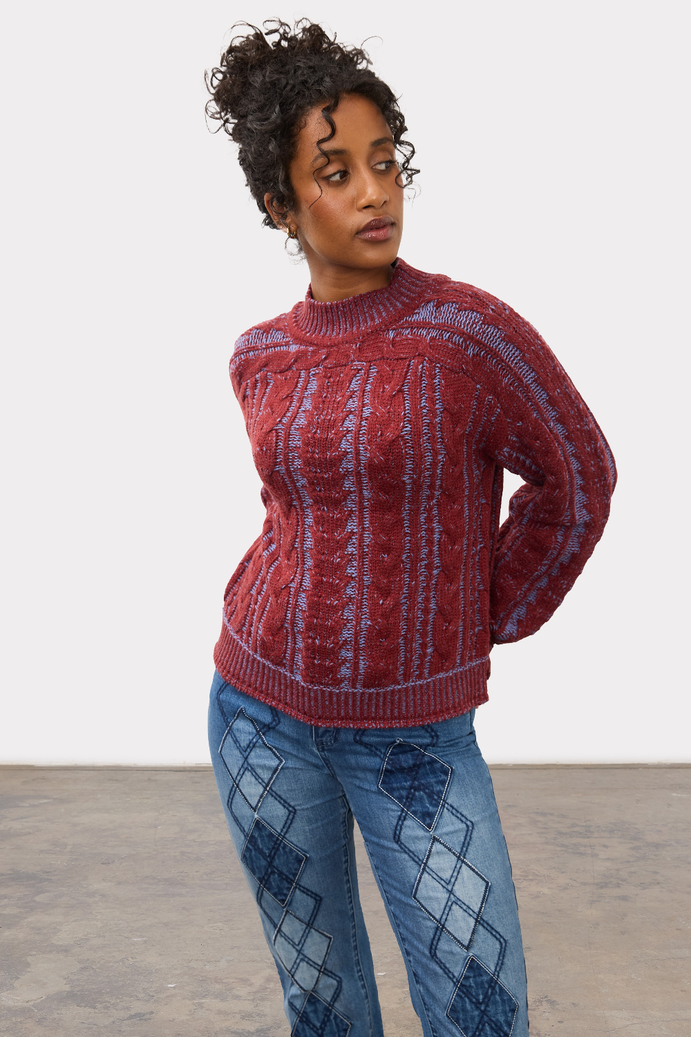 Woman wearing a red and blue mix cable knit crew neck sweater with blue jeans against a light studio backdrop.