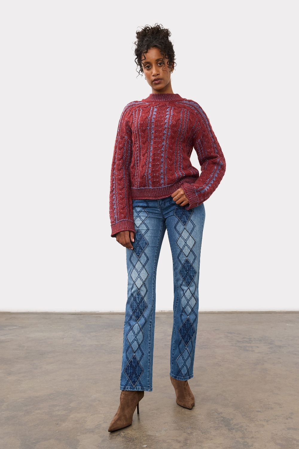 Woman wearing a red and blue mix cable knit crew neck sweater with blue jeans and brown boots against a light studio backdrop.