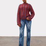 Woman wearing a red and blue mix cable knit crew neck sweater with blue jeans and brown boots against a light studio backdrop.