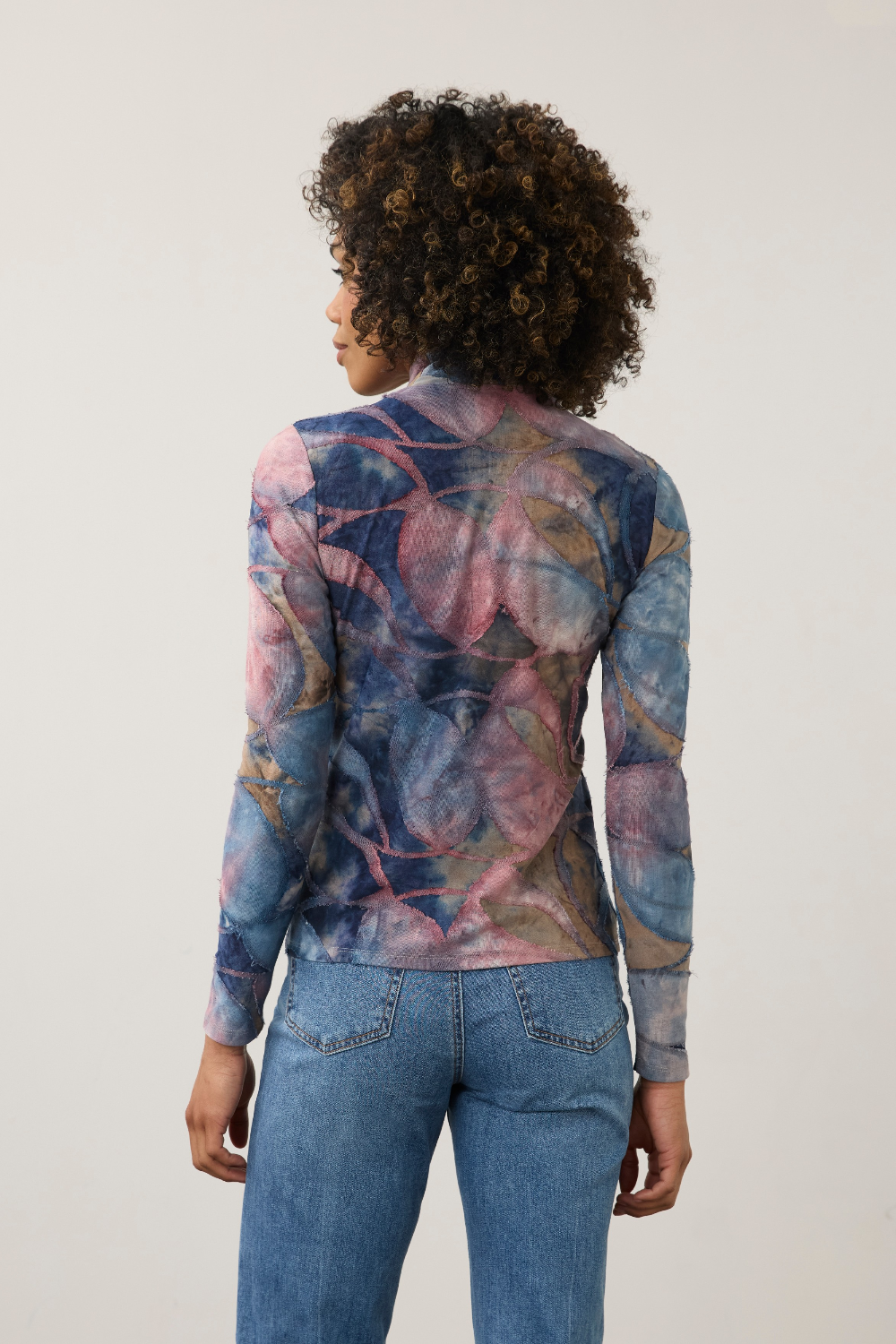 Women, shown from the back, modelling a multi coloured mock neck top with blue jeans photographed against a light background.