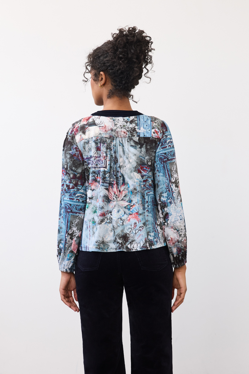Woman, shown from the back, wearing a patterned top with dark pants on a neutral background.