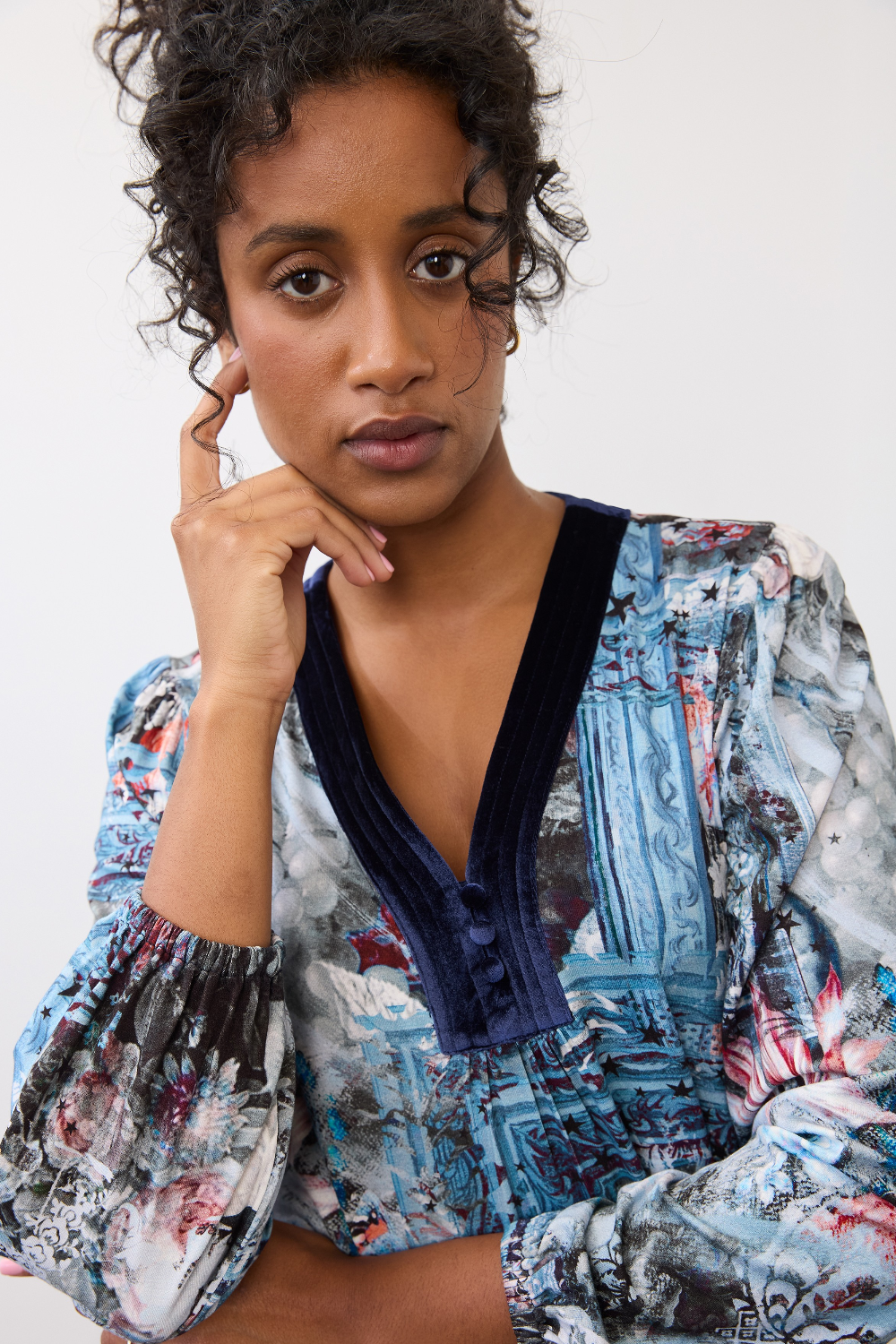 Woman wearing a patterned top, with velvet looking trim on the v-neck, on a neutral background.
