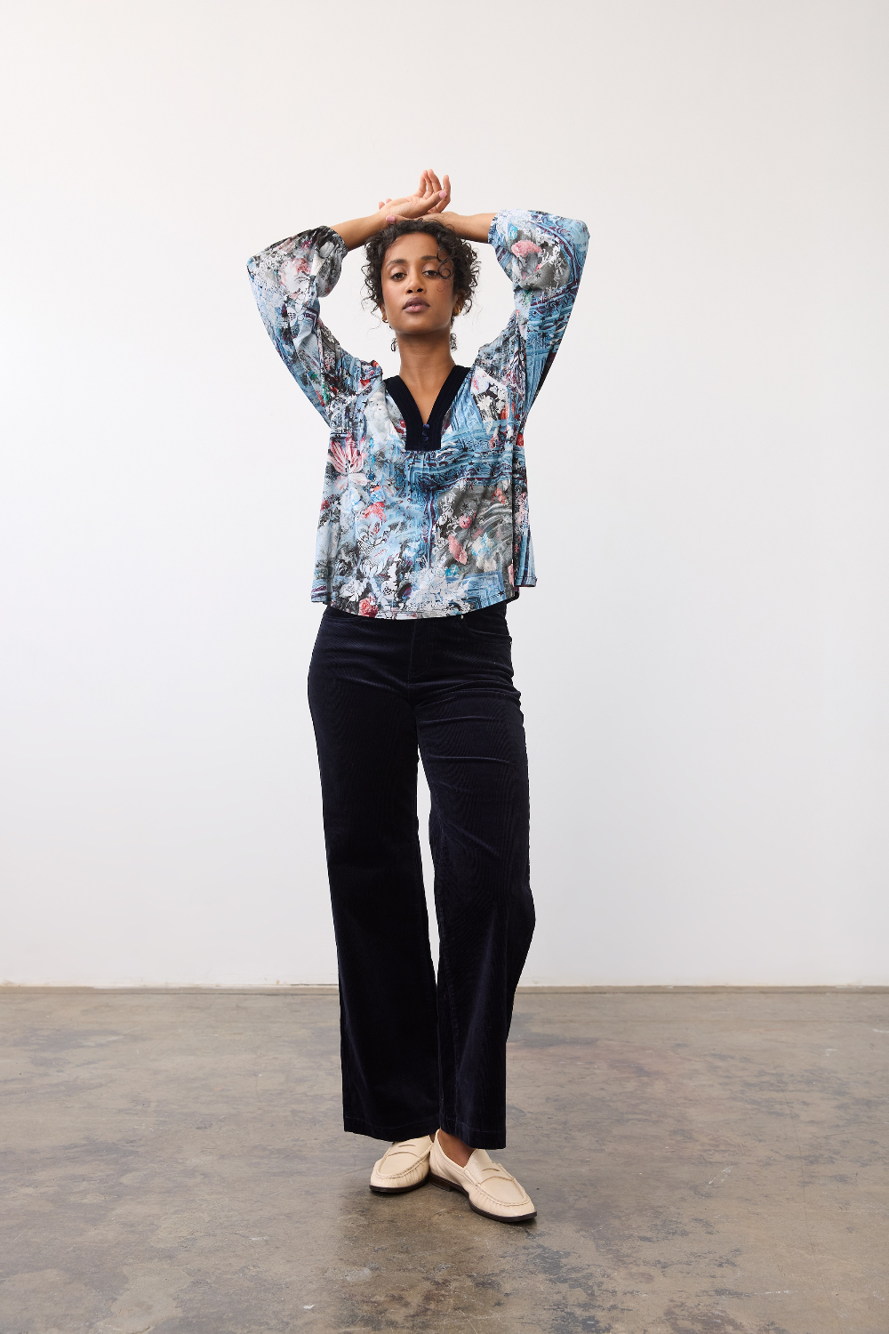 Woman wearing a patterned top, with velvet looking trim on the v-neck, with dark pants and light shoes on a neutral background.