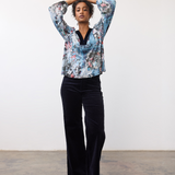 Woman wearing a patterned top, with velvet looking trim on the v-neck, with dark pants and light shoes on a neutral background.