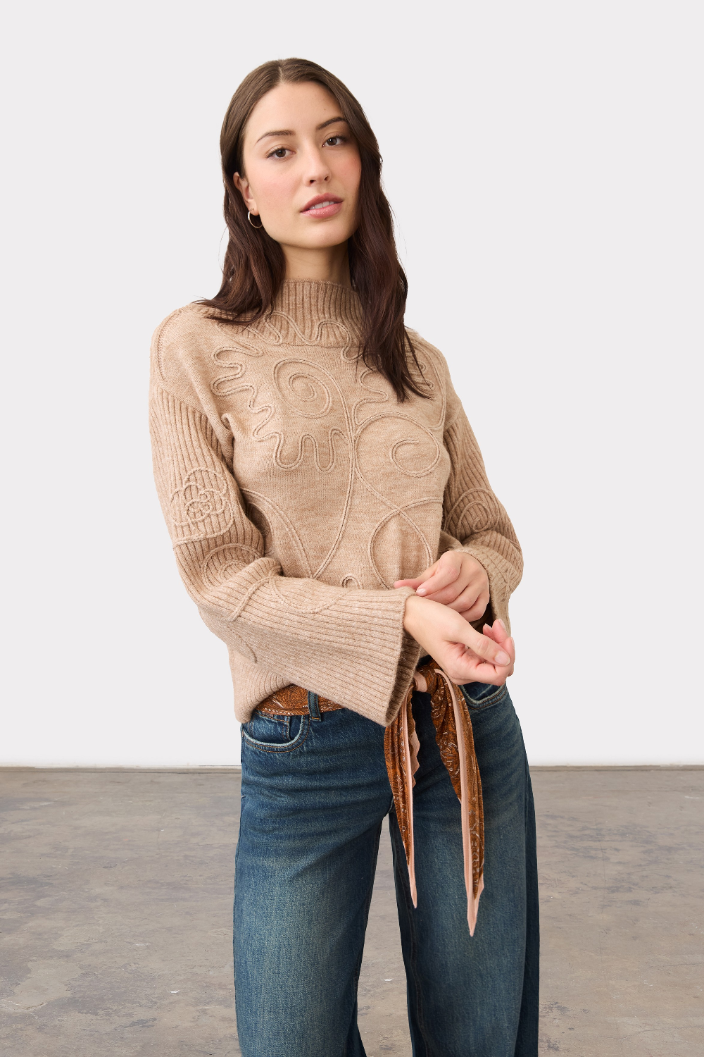 Woman modelling an embellished tan coloured sweater with belted blue jeans photographed against a light background.