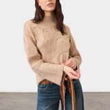 Woman modelling an embellished tan coloured sweater with belted blue jeans photographed against a light background.