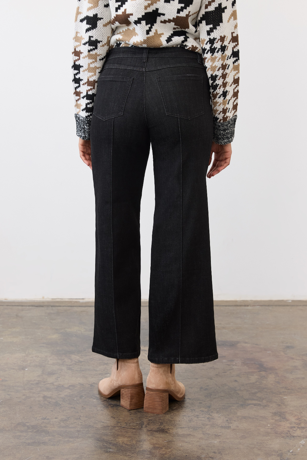 Person, shown from the back, wearing black ankle length jeans with a pin tuck down centre of the leg, with a black and tan houndstooth print sweater and tan ankle boots in a neutral studio backdrop.