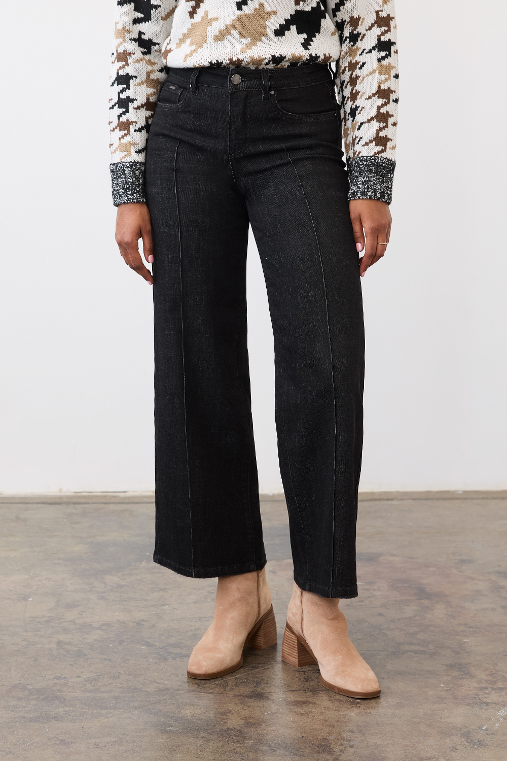 Person wearing black ankle length jeans with a pin tuck down centre of the leg, with a black and tan houndstooth print sweater and tan ankle boots in a neutral studio backdrop.