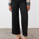 Person wearing black ankle length jeans with a pin tuck down centre of the leg, with a black and tan houndstooth print sweater and tan ankle boots in a neutral studio backdrop.