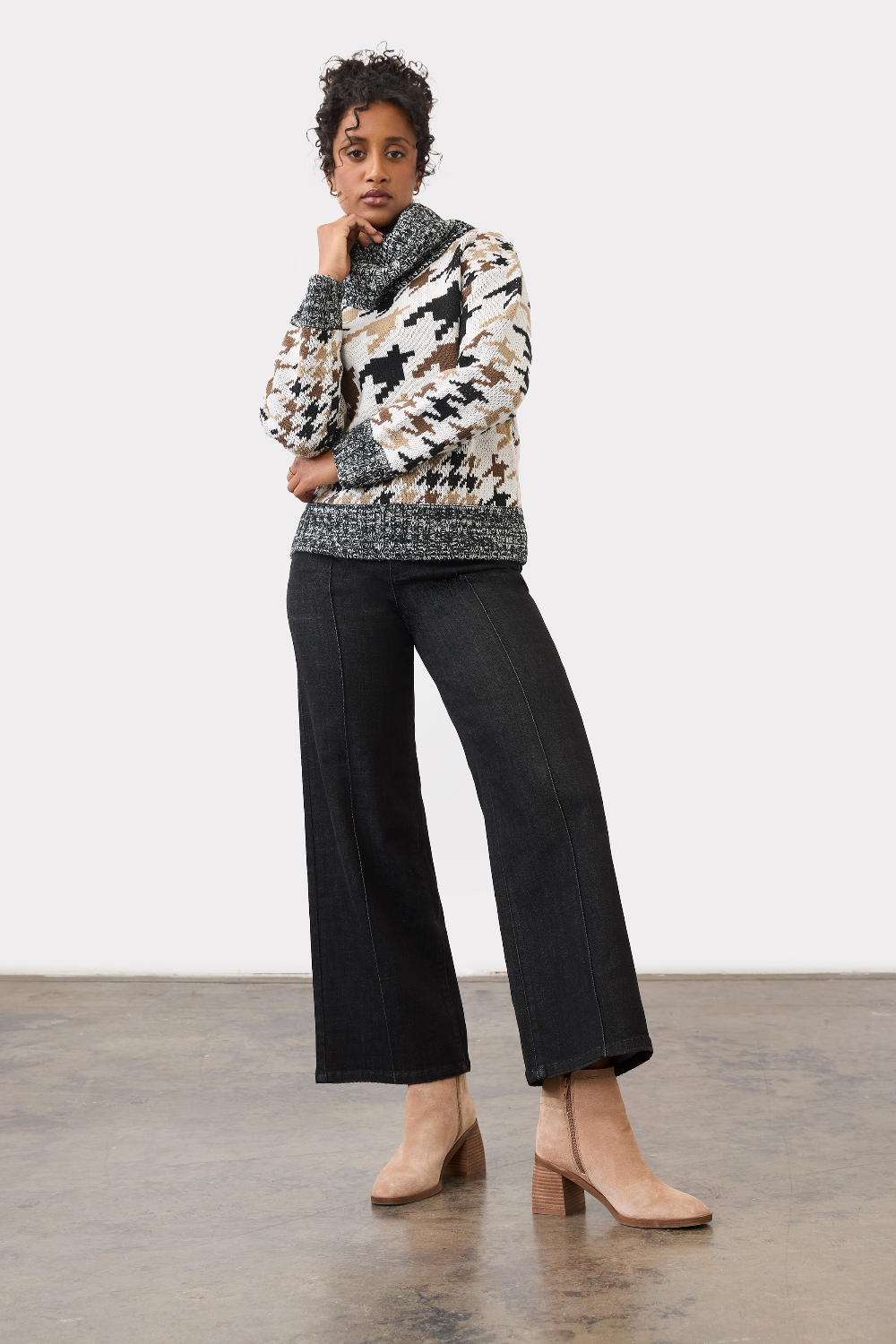 Person wearing black ankle length jeans with a pin tuck down centre of the leg, with a black and tan houndstooth print sweater and tan ankle boots in a neutral studio backdrop.
