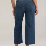 Woman, shown from behind, modelling a white tank top with wide leg cropped jeans with a pintuck and unfinished hem with black high heels against  light backdrop.