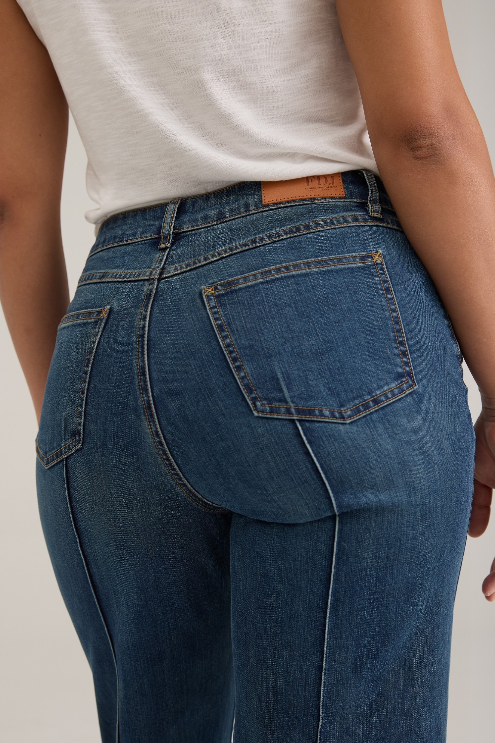 Close up of a woman, from behind, modelling a white tank top with wide leg cropped jeans with a pintuck and unfinished hem with black high heels against  light backdrop.