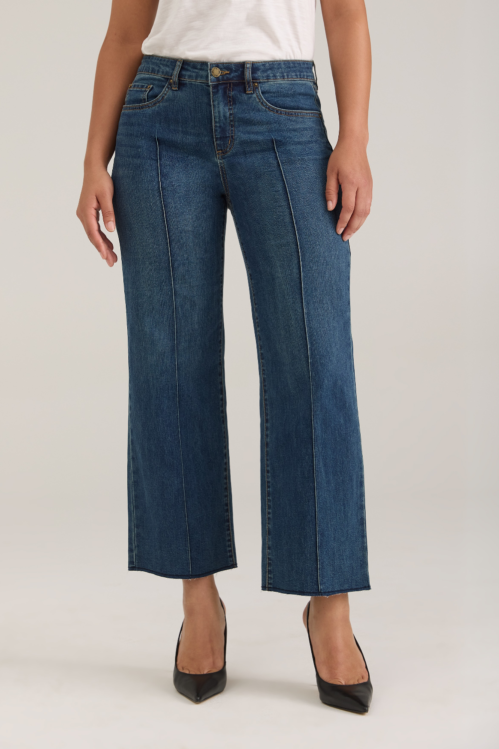 Woman modelling a white tank top with wide leg cropped jeans with a pintuck and unfinished hem with black high heels against  light backdrop.