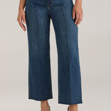 Woman modelling a white tank top with wide leg cropped jeans with a pintuck and unfinished hem with black high heels against  light backdrop.