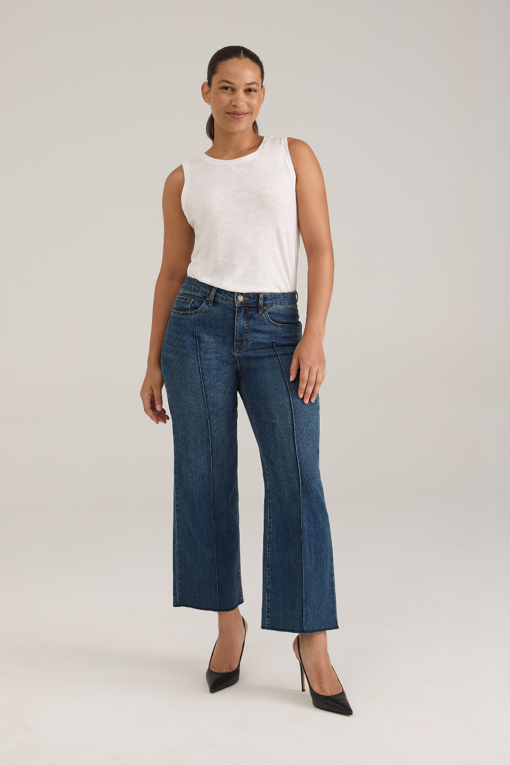 Woman modelling a white tank top with wide leg cropped jeans with a pintuck and unfinished hem with black high heels against  light backdrop.