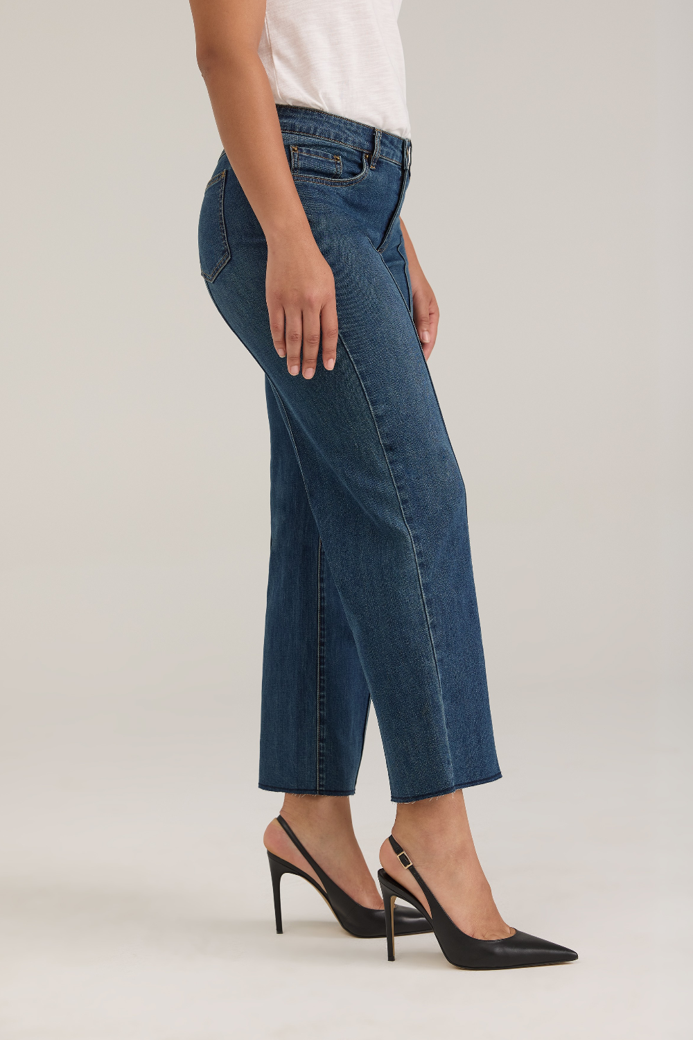 Woman modelling a white tank top with wide leg cropped jeans with a pintuck and unfinished hem with black high heels against  light backdrop.