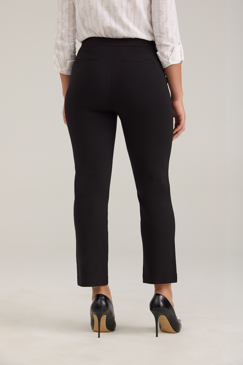 Rear view of a model wearing black cropped trousers, a white patterned blouse, and black high heels against a light gray studio background.