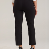 Rear view of a model wearing black cropped trousers, a white patterned blouse, and black high heels against a light gray studio background.