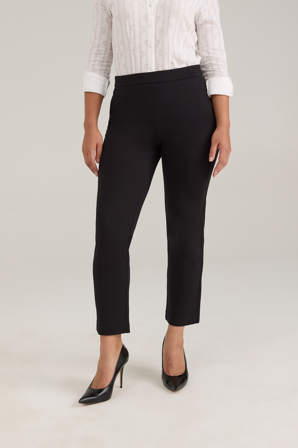 A model wearing black cropped trousers, a white textured blouse, and black high heels, standing against a light gray studio background.