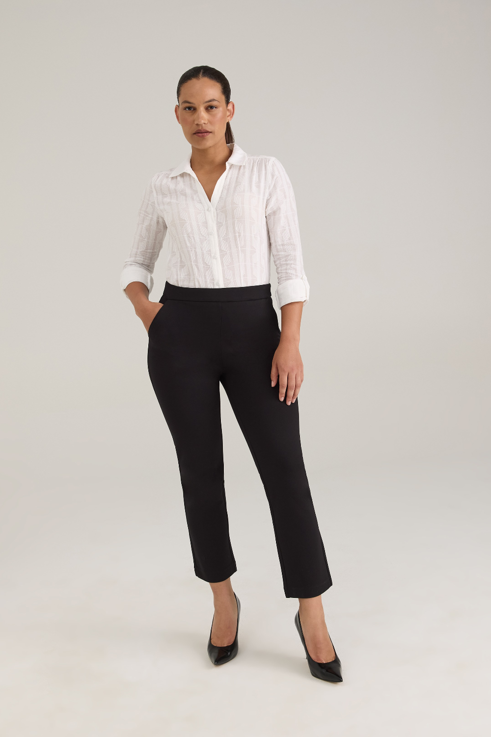 A woman modeling a white textured button-up blouse and black cropped trousers with black pointed-toe heels, standing against a light gray studio background.