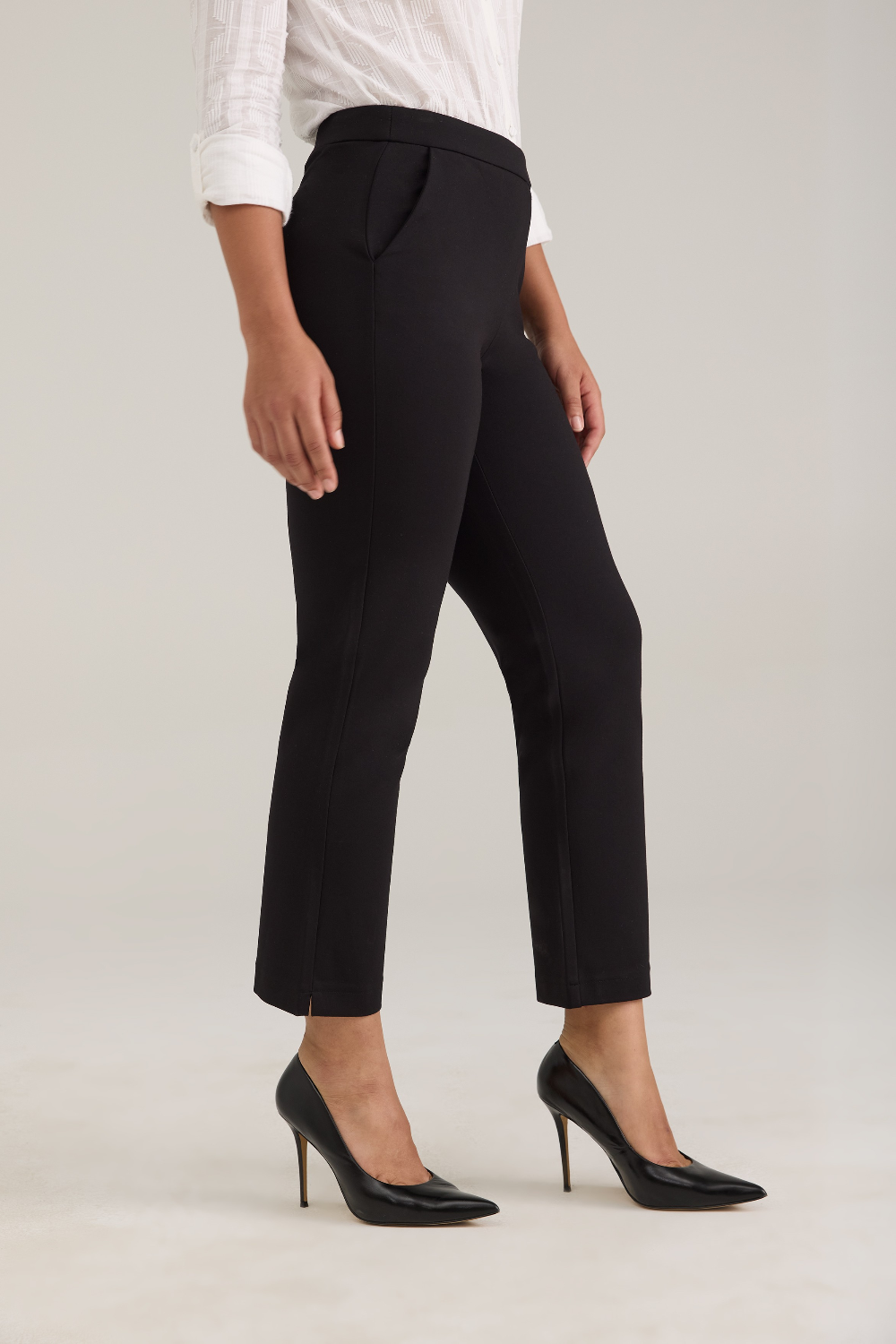 A model wearing black cropped ankle-length trousers with a white blouse and black pointed-toe high heels, shown from the waist down against a light gray studio background.