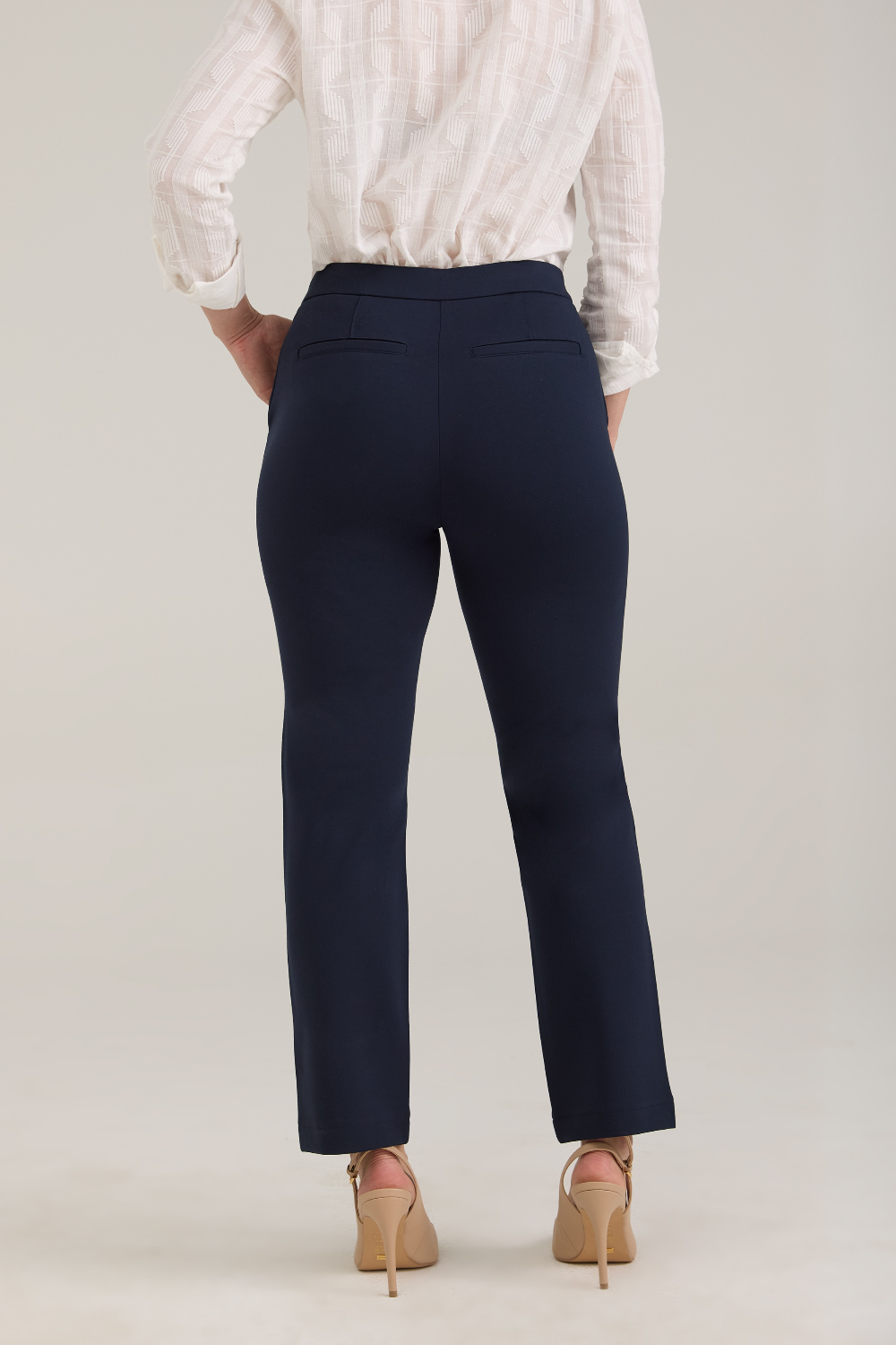 A woman, shown from the back, modeling a white textured button-up blouse and navy cropped trousers with tan pointed-toe heels, standing against a light gray studio background.