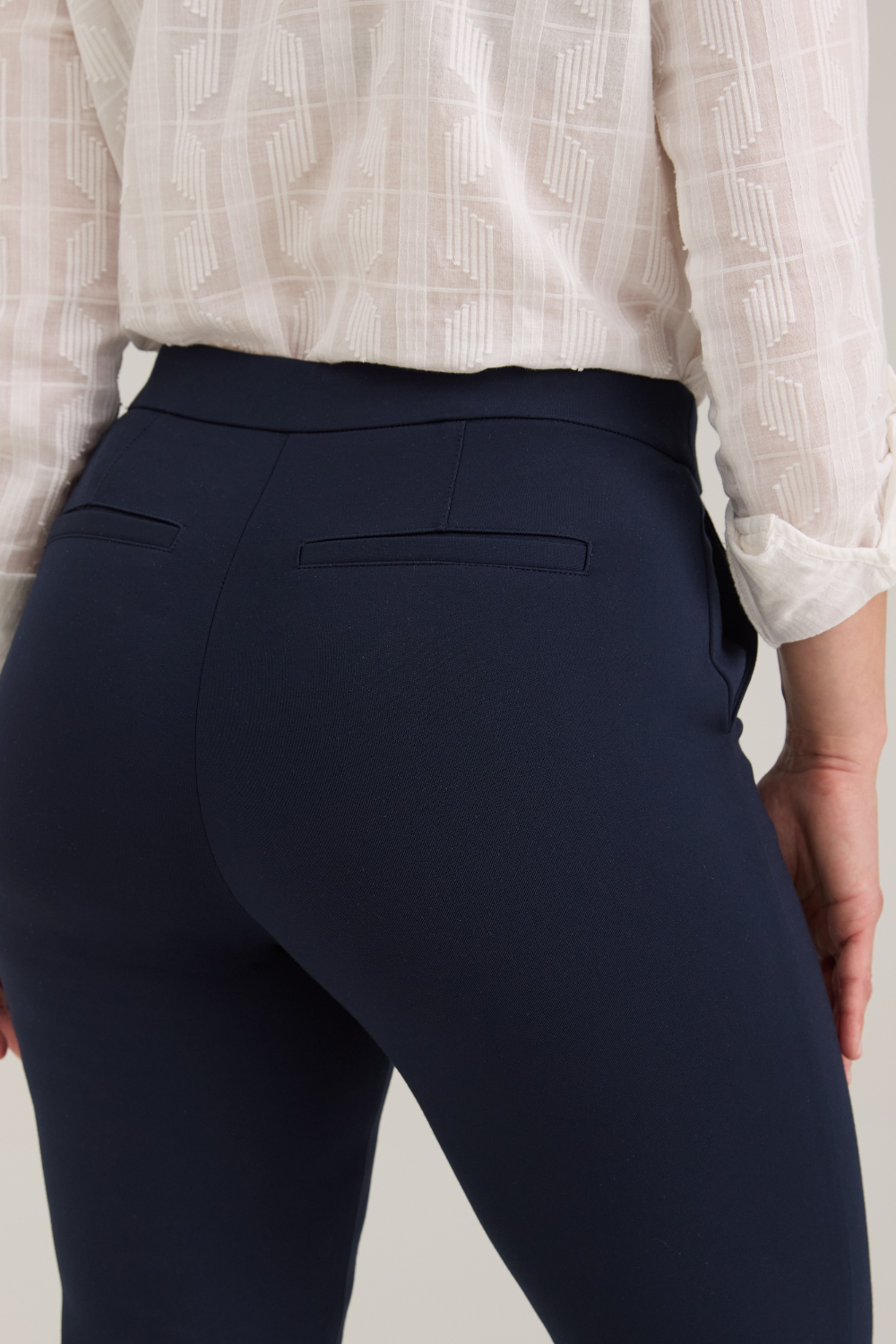 Close up of a woman, shown from the back, modeling a white textured button-up blouse and navy cropped trousers standing against a light gray studio background.