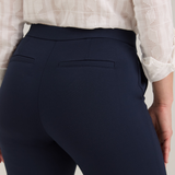 Close up of a woman, shown from the back, modeling a white textured button-up blouse and navy cropped trousers standing against a light gray studio background.