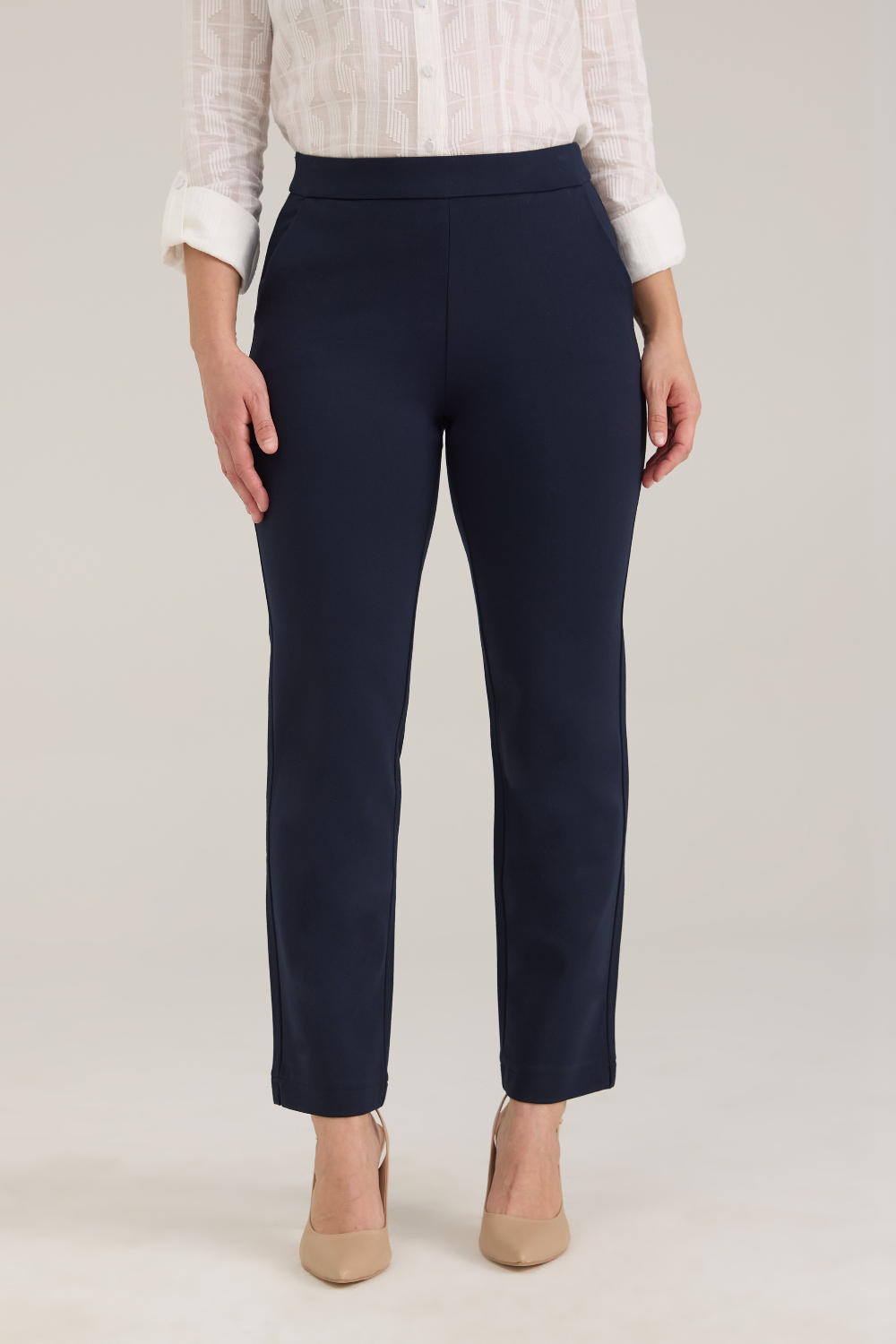 A woman modeling a white textured button-up blouse and navy cropped trousers with tan pointed-toe heels, standing against a light gray studio background.