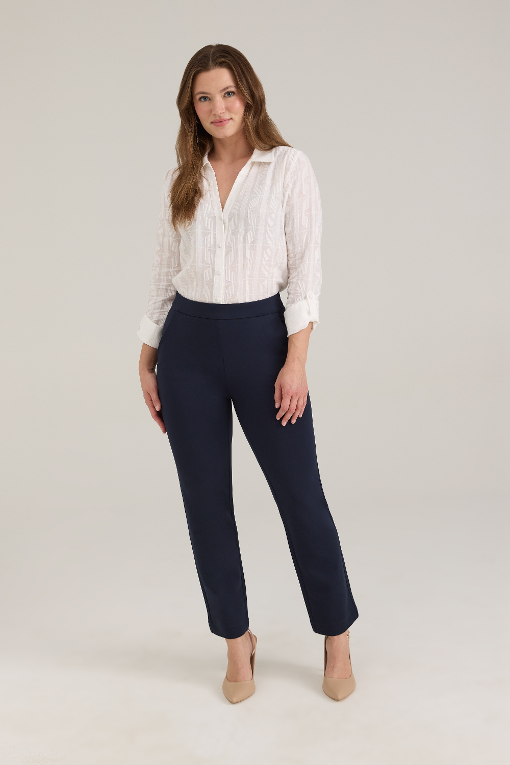 A woman modeling a white textured button-up blouse and navy cropped trousers with tan pointed-toe heels, standing against a light gray studio background.