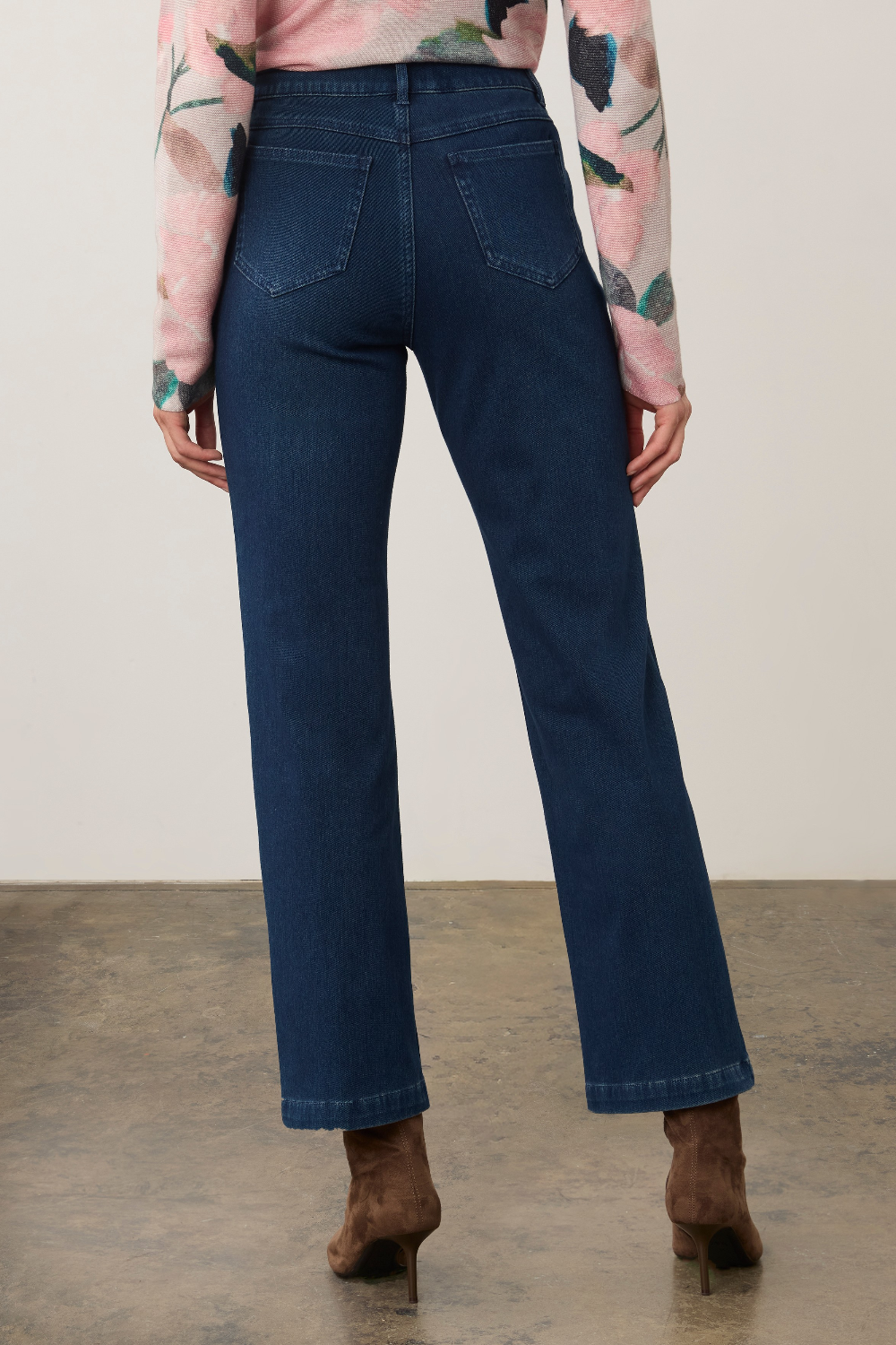 A model shown from the back wearing dark blue straight-leg jeans, a pink floral long-sleeve top, and brown suede ankle boots, standing against a neutral studio background.