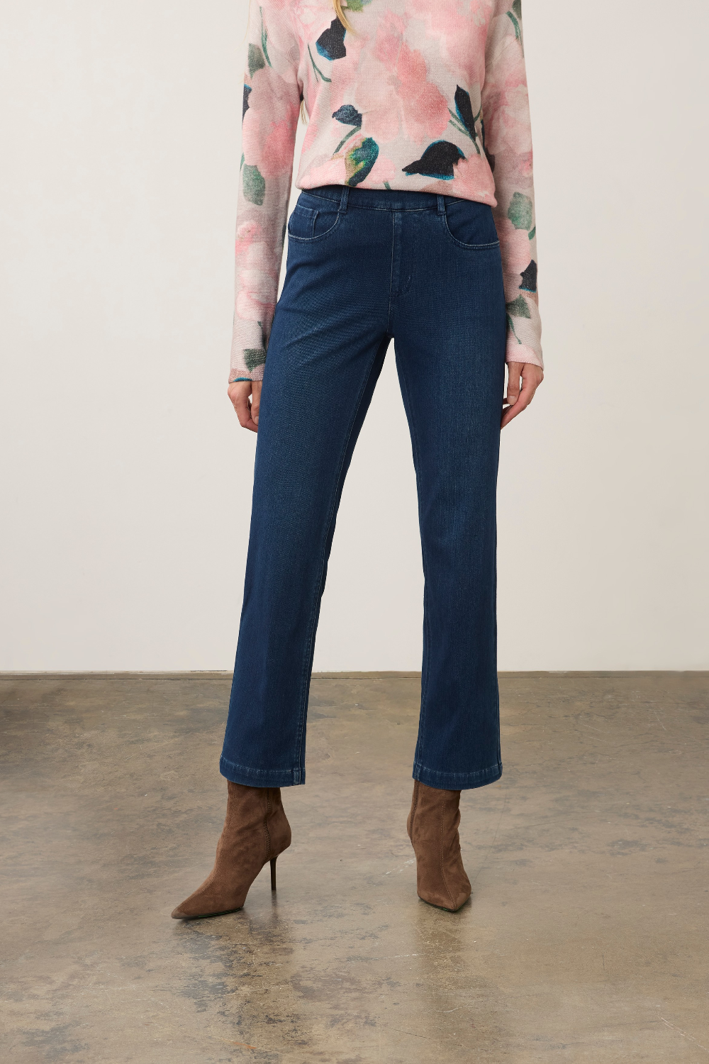 A model is shown from the waist down wearing dark blue straight-leg jeans, a pink floral long-sleeve top, and brown pointed-toe heeled ankle boots, standing against a neutral studio backdrop.
