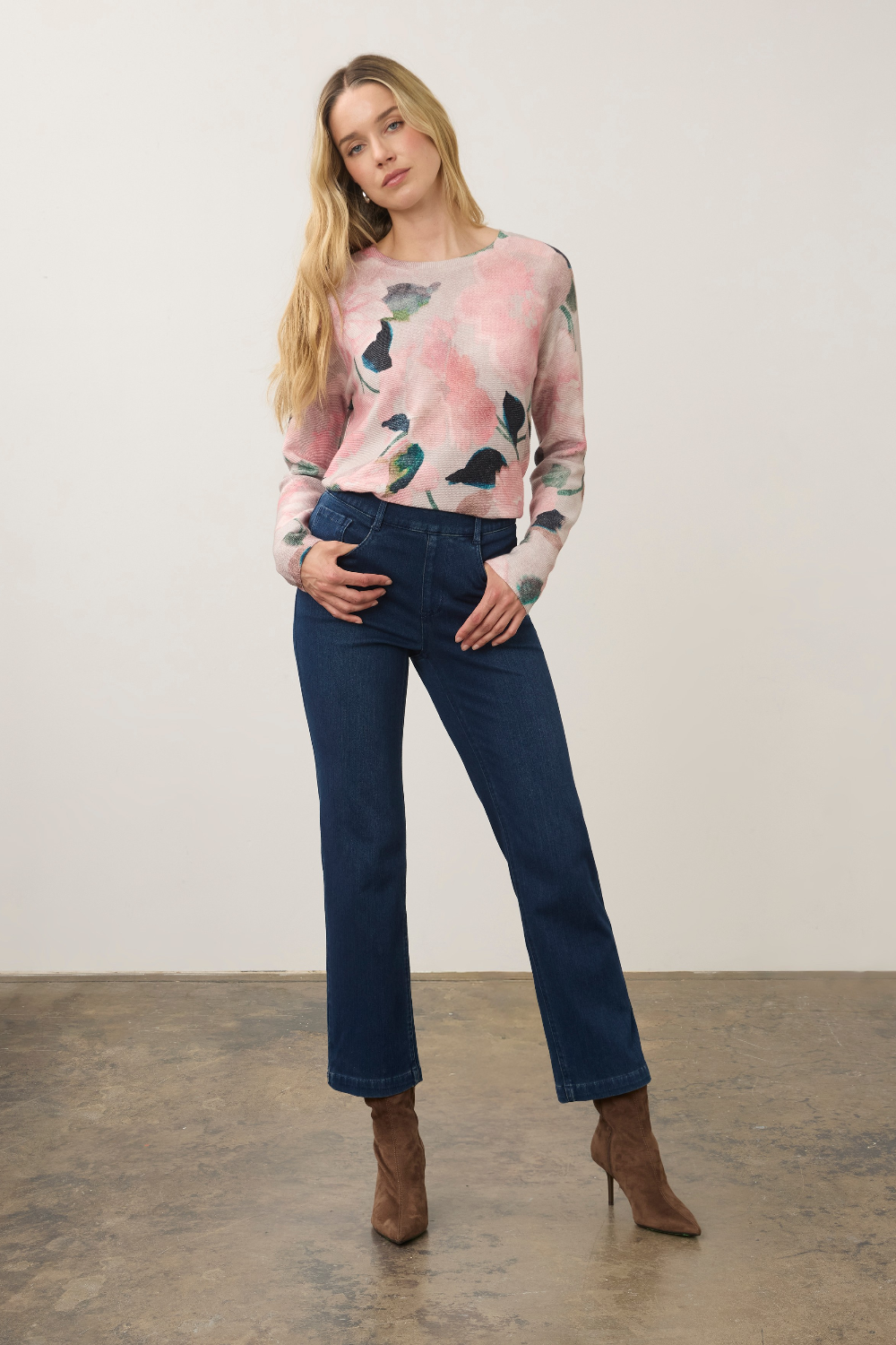 A woman modeling a light pink long-sleeve crewneck sweater with a watercolor floral pattern, paired with dark blue jeans and brown ankle boots, standing against a plain off-white wall.