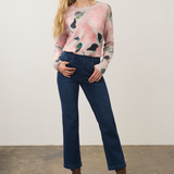 A woman modeling a light pink long-sleeve crewneck sweater with a watercolor floral pattern, paired with dark blue jeans and brown ankle boots, standing against a plain off-white wall.