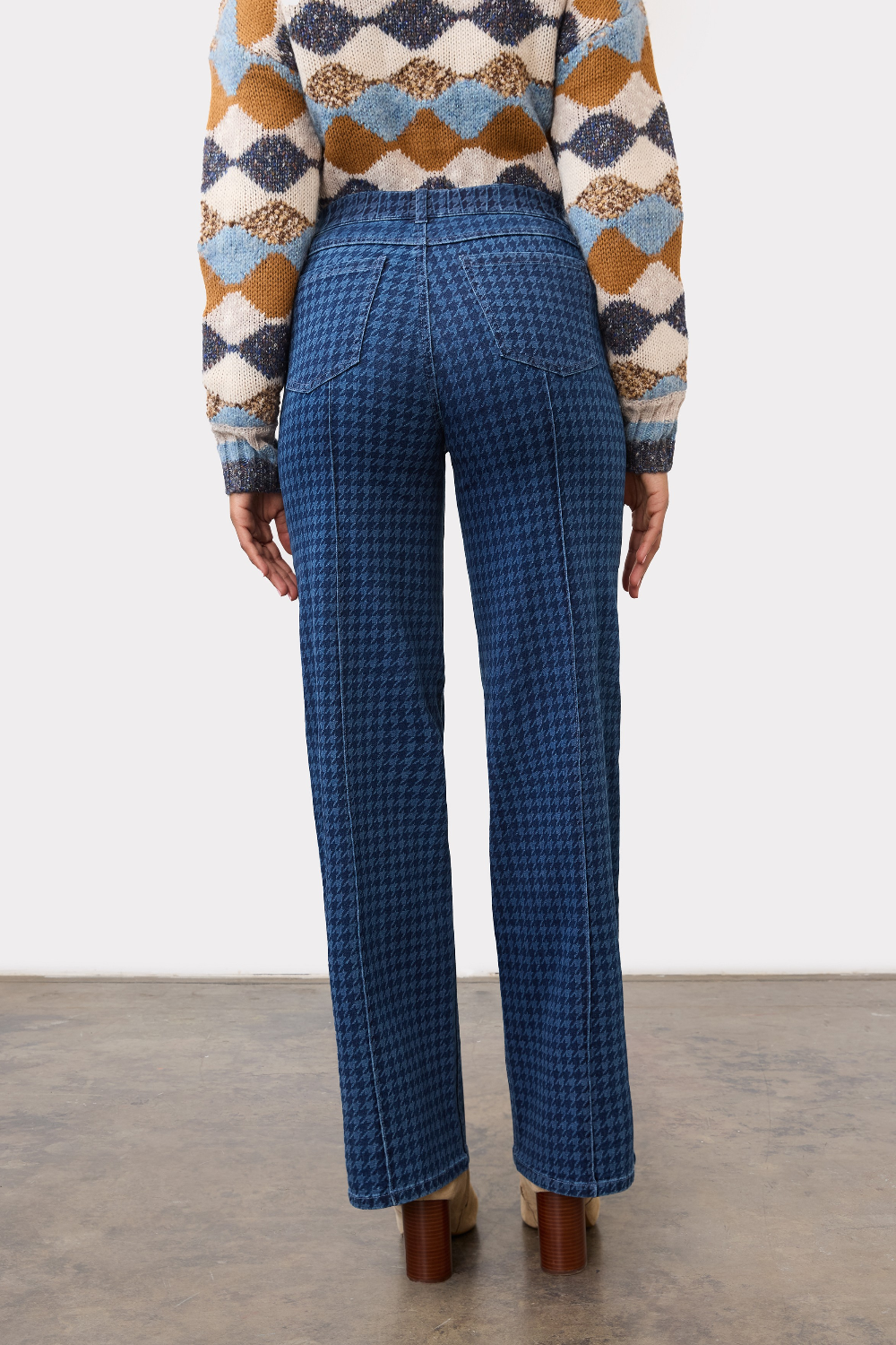 Person, shown from the back, wearing a tone on tone blue houndstooth straight leg jean, with a pintuck, with a diamond print sweater and brown boots on a neutral background.