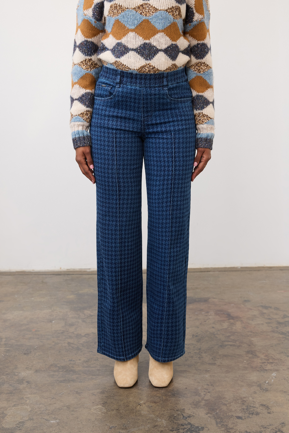 Person wearing a tone on tone blue houndstooth pull on straight leg jean, with a centre pintuck, with a diamond print sweater and tan boots on a neutral background.
