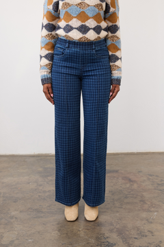 Person wearing a tone on tone blue houndstooth pull on straight leg jean, with a centre pintuck, with a diamond print sweater and tan boots on a neutral background.