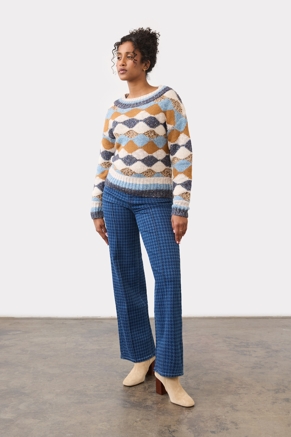 Person wearing a tone on tone blue houndstooth straight leg jean, with a centre pintuck, with a diamond print sweater and tan boots on a neutral background.