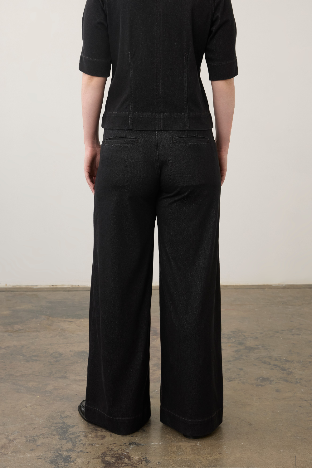 Rear view of a person wearing black wide-leg trousers and a black short-sleeve top against a plain off-white wall and concrete floor.