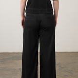 Rear view of a person wearing black wide-leg trousers and a black short-sleeve top against a plain off-white wall and concrete floor.