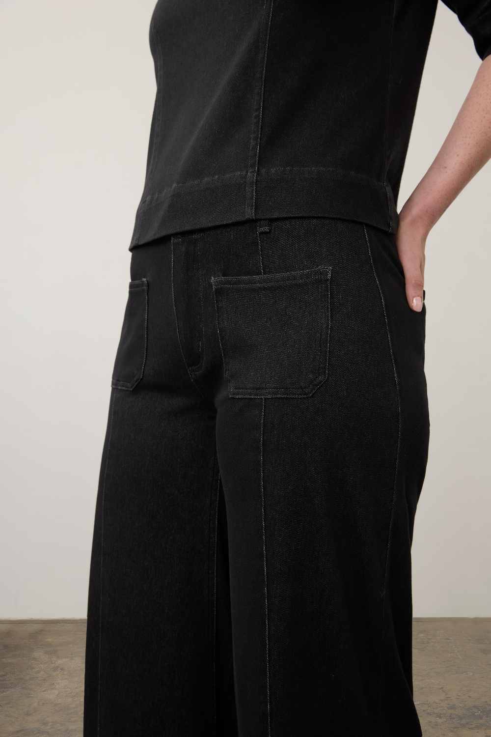 Close-up of a person wearing black wide-leg jeans with back pockets and a black top, standing against a plain light wall.