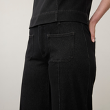 Close-up of a person wearing black wide-leg jeans with back pockets and a black top, standing against a plain light wall.